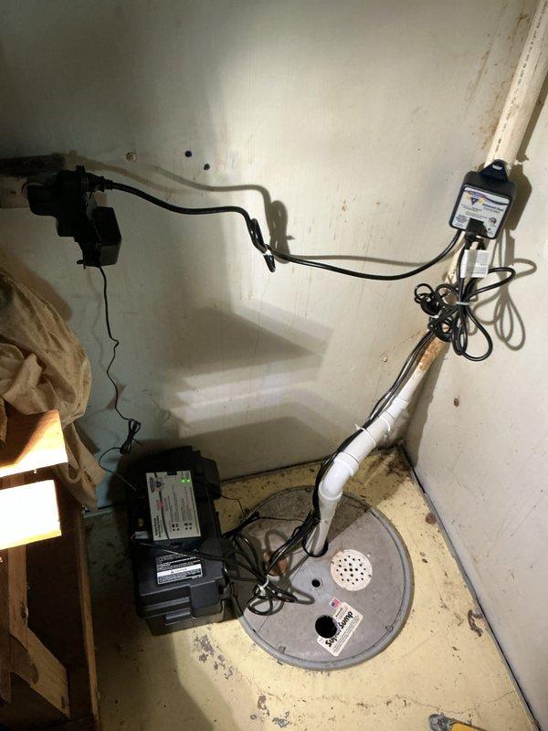 Replaced battery backup system for sump pump installation in basement utility area. New blue submersible pump installed in 18-24 inch concrete basin with white PVC discharge piping, black electrical components, and wall-mounted control switch. Parts received and system fully operational with backup power protection in place.