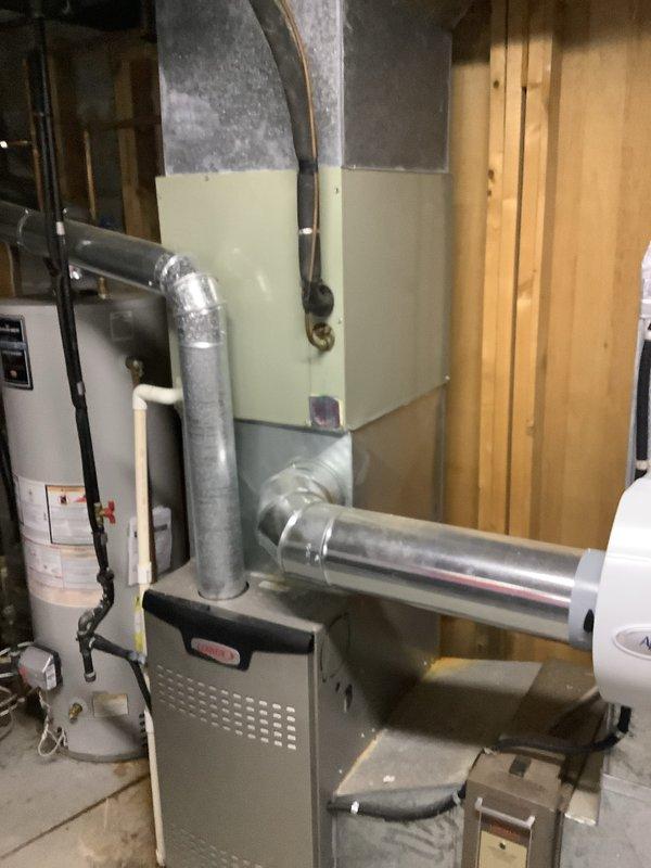 Completed installation of new Trane XE 900 air conditioning condenser unit and evaporator coil system at residential property. The outdoor condenser was positioned against the exterior foundation with proper clearances, connected to the indoor evaporator coil mounted above the existing furnace unit via refrigerant line set with black insulation. All ductwork connections were secured to the existing galvanized metal duct system, electrical connections completed, and system tested for proper operation and refrigerant charge.