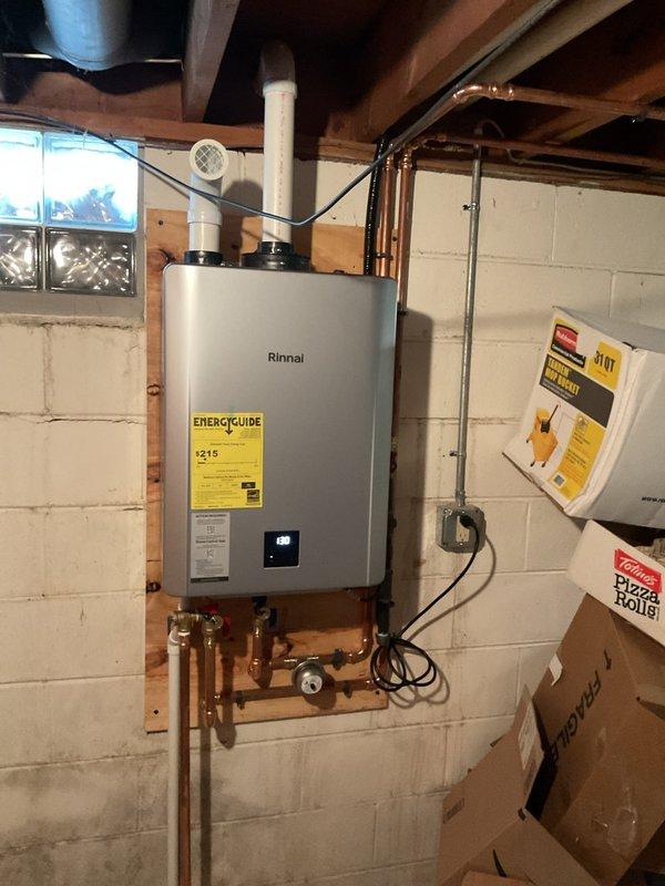 Successfully completed installation of a Rinnai RX199 tankless water heater in basement utility area. Installation included mounting the unit to concrete block wall, connecting copper supply and discharge plumbing lines, installing black ventilation piping through ceiling joists with exterior PVC termination, and completing electrical connections with new dedicated outlet. Exterior venting was properly terminated through building envelope with appropriate condensate drainage configuration utilizing PVC piping with trap assembly installed in landscaped area adjacent to structure.