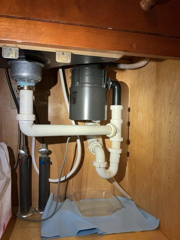 Responded to plumbing leak under kitchen sink in 94-year-old home. Upon arrival, water supply to sink was already shut off by homeowner. Inspected under-sink plumbing assembly including white PVC drain lines, P-trap configuration, garbage disposal unit, and supply connections. Identified source of leak and assessed condition of existing plumbing components. Documented cabinet interior setup with disposal motor unit, mounting hardware, and drain assembly connections. Placed protective cloth and catch container beneath plumbing to prevent water damage during inspection and repair work. Addressed leak issue, verified all connections were secure, and confirmed proper drainage function before restoring water supply to sink.