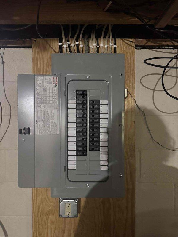 Completed 200 amp electrical service upgrade in residential basement, installing new main circuit breaker panel mounted on plywood backing board with proper conduit entry from above. Installed dedicated circuit for tankless water heater system as part of the service upgrade. Added convenience duplex receptacle outlet below main panel for utility access, with all wiring properly secured and routed through conduit per code requirements.
