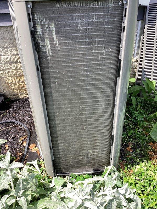 Completed routine maintenance visit on a 2-year-old residential air conditioning system for MVP customer. Upon inspection, found significant accumulation of dirt, dust, and debris on the outdoor condenser coil fins and protective grille, which can restrict airflow and reduce system efficiency. Documented unit condition with photographs showing the extent of contamination on both the heat exchanger panel and exterior mesh components.
