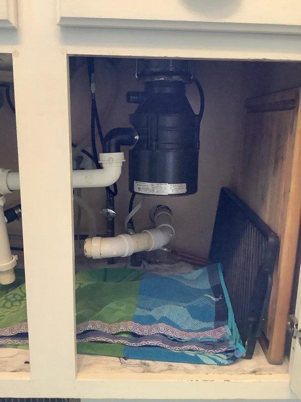 Responded to service call for clogged kitchen sink and leaking garbage disposal. Upon inspection, discovered improper sealing at disposal mounting assembly, consistent with faulty previous installation. Assessed plumbing configuration including P-trap, drain lines, and disposal connections. Placed absorbent materials beneath unit to contain active leak and protect cabinet interior. Documented existing conditions showing garbage disposal unit, PVC drainage system, and compromised seal points requiring correction.