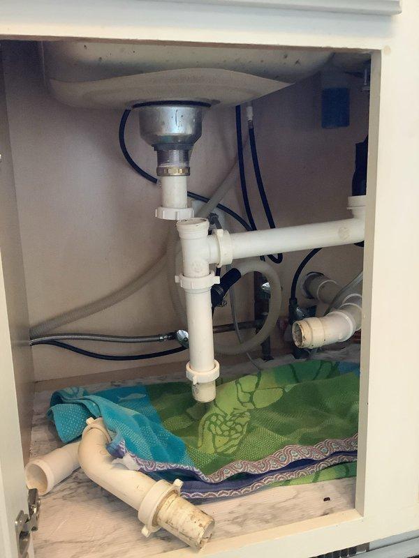 Responded to service call for clogged kitchen sink and leaking garbage disposal. Upon inspection, discovered improper sealing at disposal mounting assembly, consistent with faulty previous installation. Assessed plumbing configuration including P-trap, drain lines, and disposal connections. Placed absorbent materials beneath unit to contain active leak and protect cabinet interior. Documented existing conditions showing garbage disposal unit, PVC drainage system, and compromised seal points requiring correction.