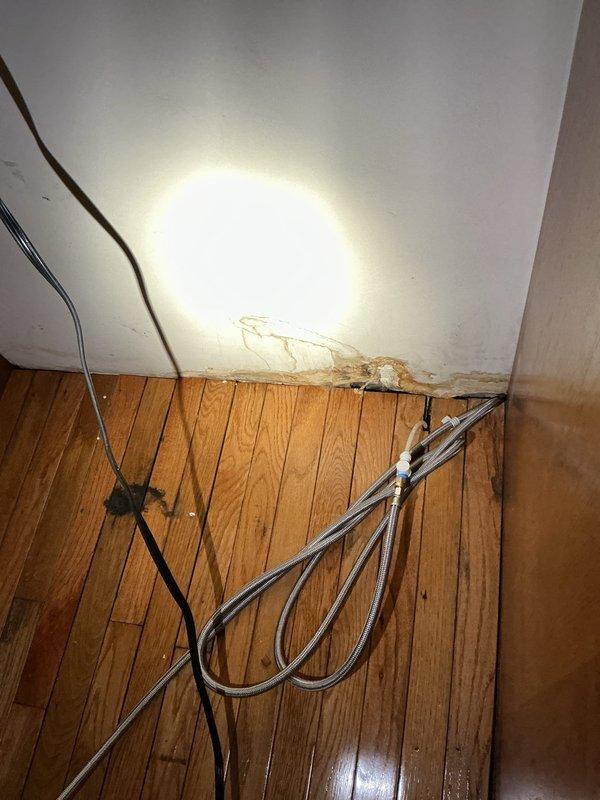 Responded to water leak in basement with no water availability during business hours. Upon inspection, identified active water damage on ceiling and walls with visible brown discoloration, peeling paint, and moisture infiltration at wall-floor junction. Documented extensive water staining along baseboards and deterioration of wall materials with cracking and flaking present. Located flexible supply line with connector valve running along affected area. Photographed damage extent showing compromised structural finishes on oak hardwood flooring and drywall surfaces for assessment and repair planning.