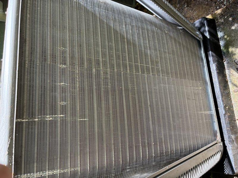 Completed routine HVAC maintenance visit for cooling system. Upon inspection, found condenser coil fins with multiple areas of damage including bent and compressed sections across horizontal bands of the heat exchanger surface. Coil exhibited heavy accumulation of dirt, dust, and outdoor debris throughout the fin surfaces, significantly impacting airflow efficiency. Documented current condition of unit with photographs showing soiled fins, debris buildup, and mechanical components including visible capacitor and wiring connections. Unit remains structurally intact despite operational wear and environmental contamination from surrounding vegetation and leaf debris.