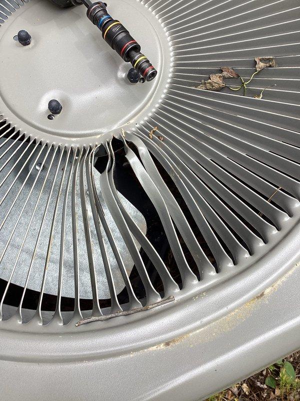 Responded to service call for outdoor condenser unit damaged by fallen tree limb that punctured the top housing. Upon inspection, found the 3-4 year old unit sustained impact damage to the upper cabinet panel, though the fan assembly, motor housing, and condenser coil fins appeared intact and undamaged. Documented unit condition with photographs showing the circular fan blade assembly, central motor housing with mounting hardware, and surrounding condenser coils all in functional condition despite debris accumulation on top of the unit from the incident.