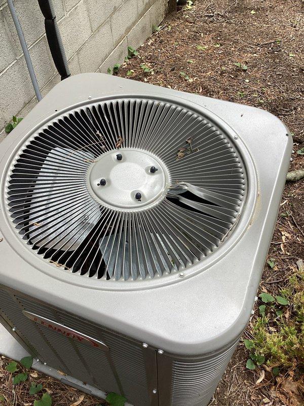 Responded to service call for outdoor condenser unit damaged by fallen tree limb that punctured the top housing. Upon inspection, found the 3-4 year old unit sustained impact damage to the upper cabinet panel, though the fan assembly, motor housing, and condenser coil fins appeared intact and undamaged. Documented unit condition with photographs showing the circular fan blade assembly, central motor housing with mounting hardware, and surrounding condenser coils all in functional condition despite debris accumulation on top of the unit from the incident.