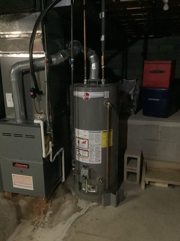 Conducted on-site assessment of existing residential tanked water heater system to provide replacement options. Inspected current gray cylindrical water heater unit located in basement utility area alongside furnace equipment. Documented water heater condition, noting visible moisture staining and discoloration on concrete floor surrounding base of unit, indicating potential minor leakage or condensation issues. Evaluated existing copper supply and distribution piping configuration extending from top of unit. Assessed installation space constraints, clearances, and existing plumbing connections to determine feasibility for both traditional tanked and tankless water heater replacement options. Photographed equipment labels, specifications, and surrounding mechanical systems including overhead plumbing runs and utility connections to support accurate quote preparation for customer's requested replacement alternatives.