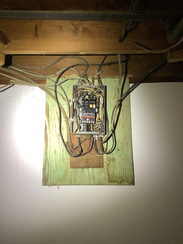 Technician responded to evaluate electrical panel and associated repairs in basement utility area. Upon inspection, found older-style electrical panel with exposed breakers and wiring connections requiring assessment. Panel was mounted on wooden backing board with multiple circuit breakers and various black and gray cables entering from multiple directions. Documented current panel condition and wiring configuration through photographs for repair recommendations. Additionally inspected adjacent utility space with exposed framing and concrete block walls, noting vertical conduit installation secured with mounting brackets. Evaluated overall electrical infrastructure in unfinished basement area to determine necessary repairs and code compliance requirements for panel and associated electrical systems.