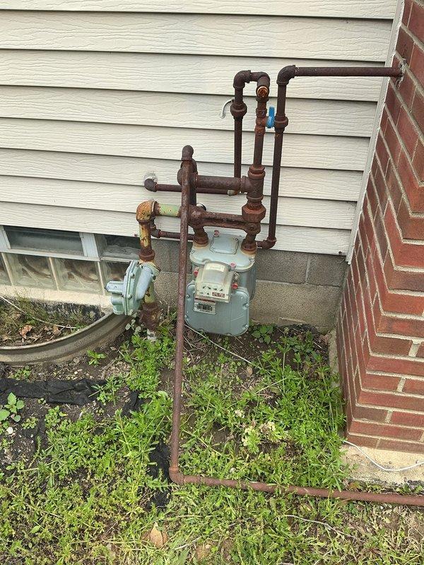 Responded to service call for gas line connection estimate to accommodate new gas heater installation. Upon arrival, conducted thorough inspection of existing exterior gas meter and associated piping system mounted on vinyl siding near foundation. Documented current condition of gas infrastructure, noting significant rust and corrosion present on multiple pipe fittings, valves, and connection points throughout the aged installation. Assessed routing requirements and connection feasibility from existing gas meter to proposed heater location. Photographed existing gas meter assembly, piping configuration including vertical and horizontal runs, shut-off valve placement, and overall system condition to support estimate preparation. Evaluated structural considerations including proximity to brick foundation, basement window clearance, and optimal pathway for new gas line run. Determined scope of work necessary for safe and code-compliant gas line extension to support new heating equipment i