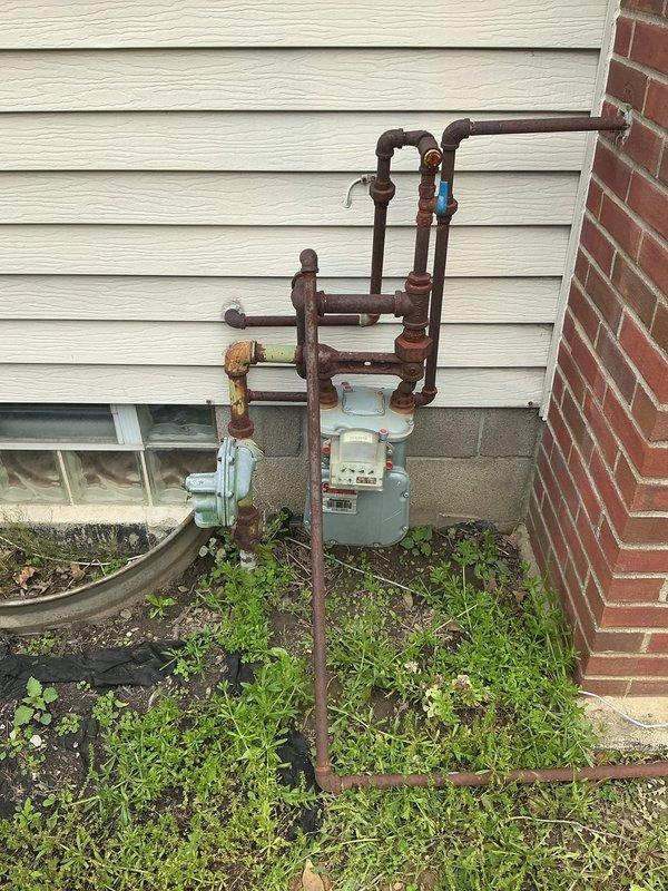 Responded to service call for gas line connection estimate to accommodate new gas heater installation. Upon arrival, conducted thorough inspection of existing exterior gas meter and associated piping system mounted on vinyl siding near foundation. Documented current condition of gas infrastructure, noting significant rust and corrosion present on multiple pipe fittings, valves, and connection points throughout the aged installation. Assessed routing requirements and connection feasibility from existing gas meter to proposed heater location. Photographed existing gas meter assembly, piping configuration including vertical and horizontal runs, shut-off valve placement, and overall system condition to support estimate preparation. Evaluated structural considerations including proximity to brick foundation, basement window clearance, and optimal pathway for new gas line run. Determined scope of work necessary for safe and code-compliant gas line extension to support new heating equipment i