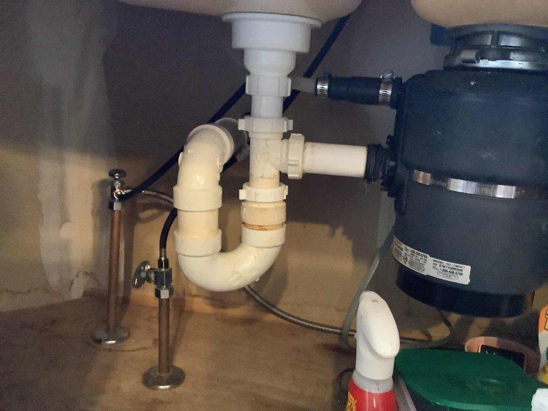 Responded to service call for reported leak from rear faucet causing water intrusion into basement area. Upon arrival, conducted thorough inspection of kitchen sink plumbing system and underlying basement structure. Examined under-sink cabinet installation including PVC drain assembly, P-trap configuration, garbage disposal unit, and flexible water supply lines. Inspected basement ceiling area directly below kitchen plumbing, identifying moisture evidence on painted wooden joist with visible stress crack and copper supply line routing. Assessed all connection points, drain components, and supply line fittings for signs of active leakage or moisture accumulation. Documented plumbing configuration and structural conditions with photographic evidence for service records.