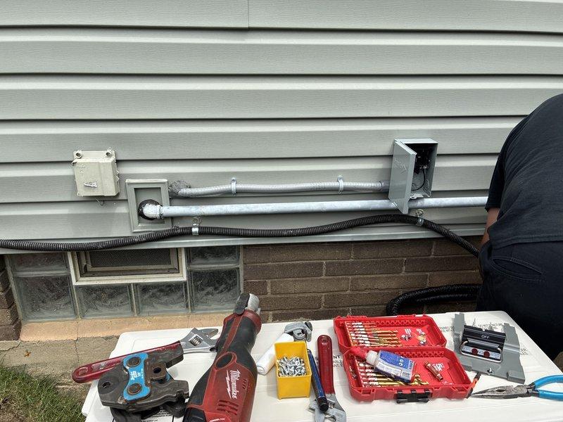 Installed new 480V AC circuit wiring with exterior conduit routing along building facade, securing dual parallel runs of rigid metal conduit with appropriate mounting hardware and weatherproof junction boxes. Completed interior basement wiring connections, routing conductors through floor joists and integrating with existing electrical infrastructure while maintaining proper clearances around glass block window installation and ventilation components.