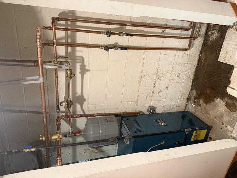 Successfully completed hot water boiler installation in basement mechanical room. Installed new blue residential boiler unit with accompanying gray expansion tank, configured with comprehensive copper piping manifold system featuring multiple zone distribution lines. System includes properly installed supply and return lines, zone valves, and all necessary connections for hydronic heating operation. Integrated new boiler with existing ductwork and HVAC infrastructure, ensuring proper venting and clearances per manufacturer specifications. All copper piping professionally soldered and secured, expansion tank properly mounted and pressurized, and system tested for leaks and proper operation. Installation meets current mechanical codes and manufacturer requirements for residential hydronic heating systems.