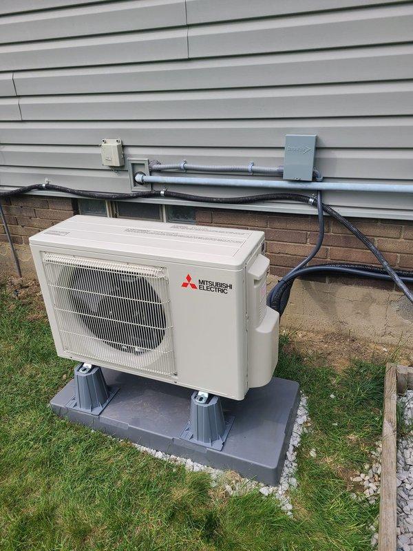 Completed installation of Mitsubishi Electric mini split system including outdoor condensing unit mounted on composite pad with proper elevation and leveling. Installed electrical conduit routing from unit through brick foundation to wall-mounted disconnect box and junction box on exterior siding, secured all components with appropriate mounting hardware and brackets. System positioned on prepared site with landscape edging and stone base for drainage and stability.