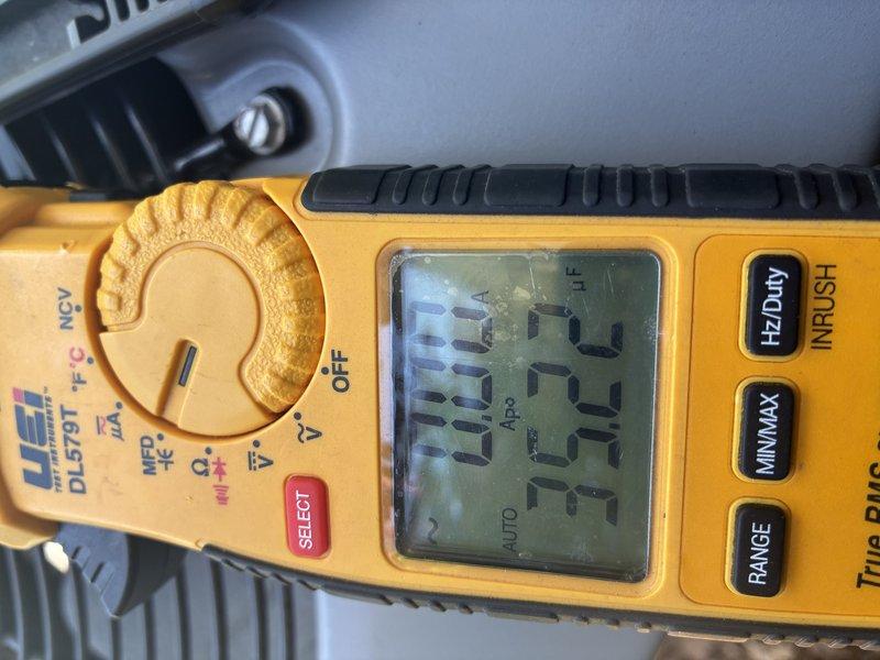 Performed comprehensive air conditioner tune-up on outdoor condensing unit. Conducted electrical diagnostics using digital multimeter to verify proper voltage and amperage readings, with measurements indicating system operating within normal parameters at approximately 50.5 volts during testing. Inspected condenser unit exterior condition, noting accumulated debris and weathering on mesh grill covering that required attention for optimal airflow and system efficiency.