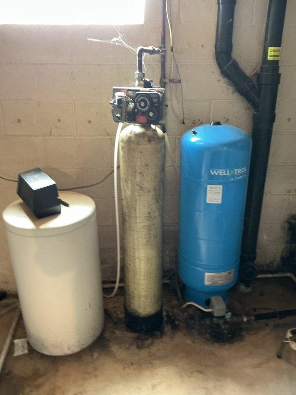 Responded to service call for water heater leaking from bottom. Upon inspection, found existing tanked water heater showing significant corrosion and deterioration on tank surface with visible rust patches and wear throughout the unit. Leak originated from bottom of tank, indicating tank failure. Documented condition of water heater and surrounding plumbing components including pressure tank, water softener, and associated piping systems. Provided assessment and quote for water heater replacement, as unit is beyond economical repair due to extent of tank corrosion and active leak from base.