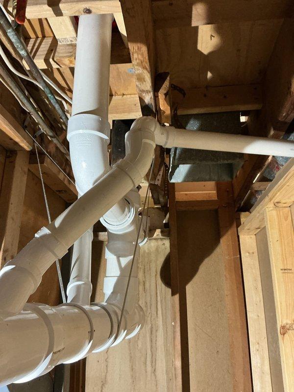 Replaced deteriorated cast iron drain lines with new PVC piping from 4-inch fernco connection to back-to-back bathroom group configuration. Removed existing cast iron drainage system and installed white PVC drain pipes with proper fittings, elbows, and couplings through wooden floor framing. Secured all piping with appropriate clamps and supports, ensuring proper pitch and alignment for optimal drainage flow. Two-person crew completed full-day installation including rough-in plumbing work through subfloor and structural framework.