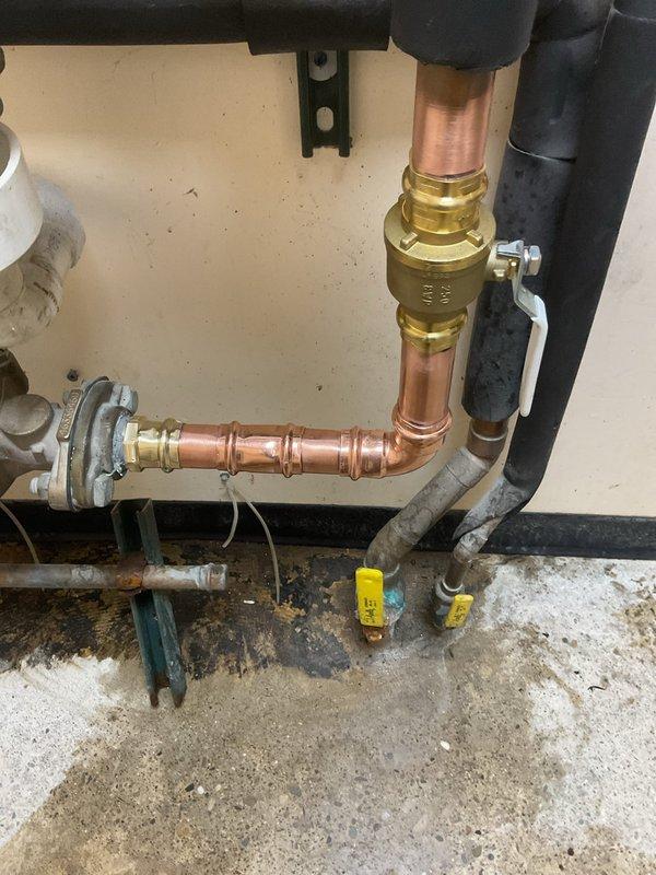 Responded to a leak at a 1 1/2 x 1 x 1 1/2 copper tee fitting located on the outlet side of the water meter. Rebuilt the configuration from the meter flange, eliminating the problematic 1-inch pipe section, installed a new brass ball valve on the vertical copper pipe run, and securely reconnected all components to the meter flange. The repair successfully resolved the leak and restored proper water flow to the system.