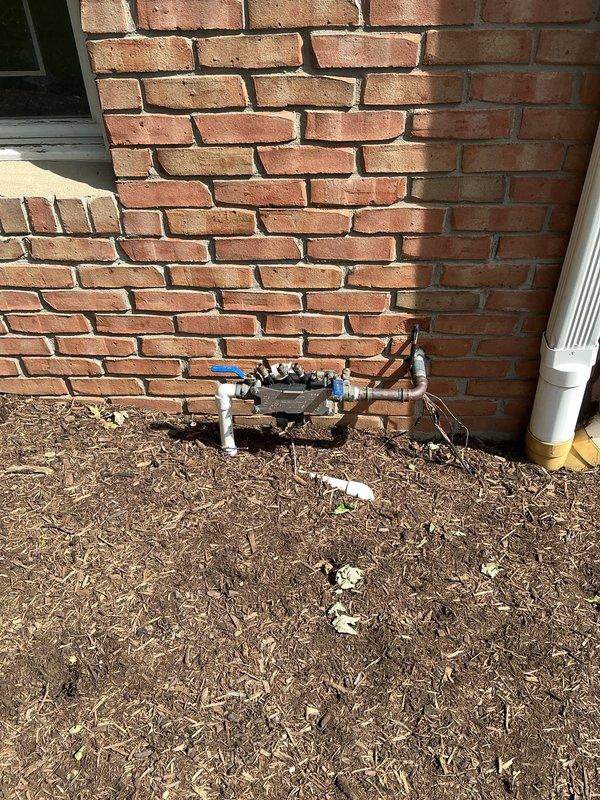 Responded to service call for irrigation backflow preventer with reported small leak. Upon inspection, identified leak at backflow prevention device mounted on exterior brick wall with white PVC piping connections. Device features multiple brass threaded fittings and blue valve handle component. Documented existing conditions with photos showing backflow assembly, manifold connections, and surrounding plumbing infrastructure for service records and future reference.