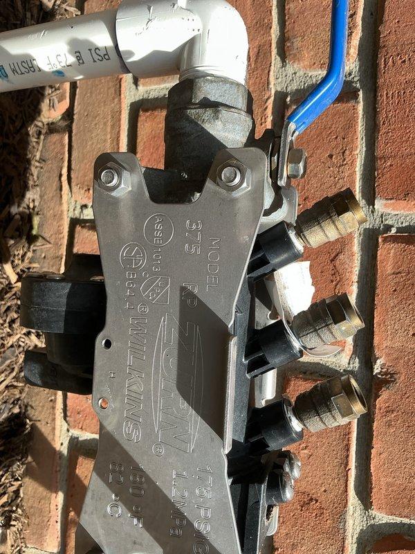 Responded to service call for irrigation backflow preventer with reported small leak. Upon inspection, identified leak at backflow prevention device mounted on exterior brick wall with white PVC piping connections. Device features multiple brass threaded fittings and blue valve handle component. Documented existing conditions with photos showing backflow assembly, manifold connections, and surrounding plumbing infrastructure for service records and future reference.
