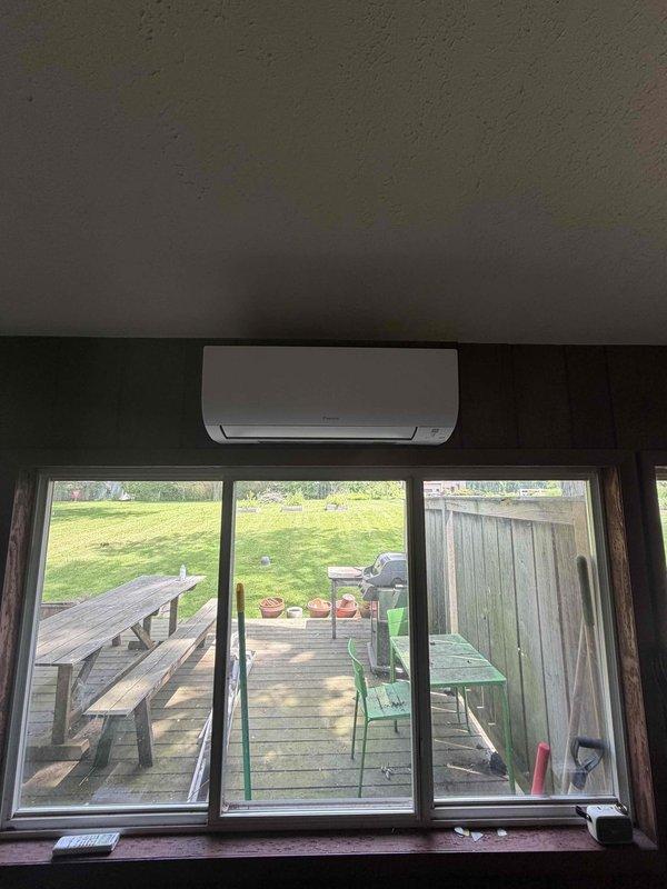 Completed installation of a single-head Daikin ductless mini-split system. Mounted indoor air handler unit on interior wall above sliding glass door, installed outdoor condenser unit on concrete pad against exterior wall with light blue siding, and ran refrigerant lines through protective conduit connecting both units. System tested and confirmed operational.