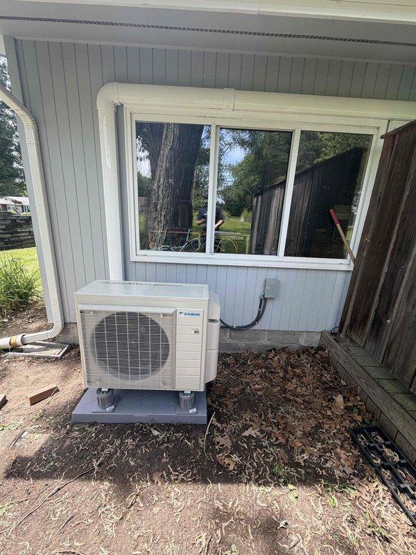 Completed installation of a single-head Daikin ductless mini-split system. Mounted indoor air handler unit on interior wall above sliding glass door, installed outdoor condenser unit on concrete pad against exterior wall with light blue siding, and ran refrigerant lines through protective conduit connecting both units. System tested and confirmed operational.