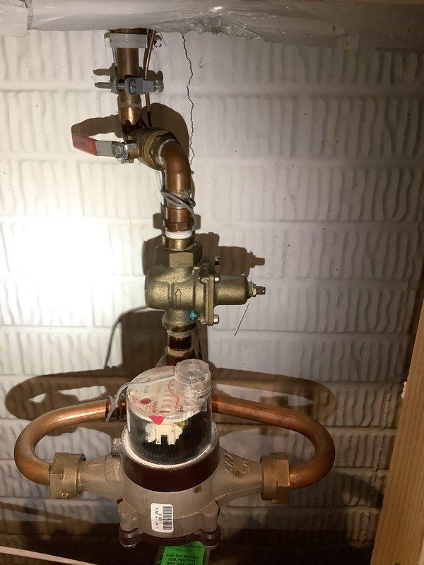 Responded to service call for knocking sound near waterlines and valve area. Upon inspection, examined main water meter assembly, shut-off valves, and associated copper piping mounted at wall. Assessed water heater installation including connected plumbing, expansion tank, and supply lines for potential sources of water hammer or loose components that could generate knocking sounds during water flow or pressure changes.
