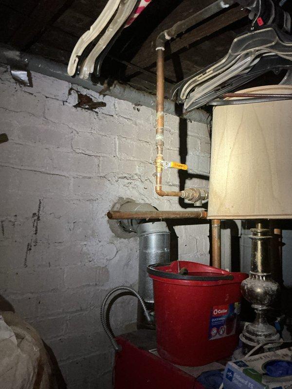 Responded to service call for 20+ year old steam boiler with missing copper piping. Upon arrival, found water to home was shut off due to incomplete piping connections. Assessed boiler system and identified missing copper supply line sections that required installation. Documented existing conditions showing water heater, associated plumbing infrastructure with brass fittings and valves, and basement utility area layout. Photographed piping connections, structural elements, and overall system configuration for repair planning and customer records.
