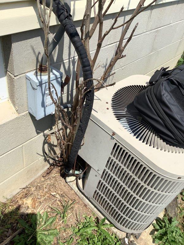 Conducted on-site evaluation of 22-year-old air conditioning system for replacement estimate. Inspected outdoor condenser unit and assessed A-coil replacement requirements. Unit showed signs of age-appropriate wear, with vegetation debris accumulation on top of condenser and overgrown plant material in immediate vicinity affecting proper airflow and maintenance access. Photographed existing equipment configuration, electrical disconnect box, and refrigerant line routing for replacement planning. Documented current system specifications and site conditions to provide accurate equipment sizing and installation scope for complete HVAC system replacement.