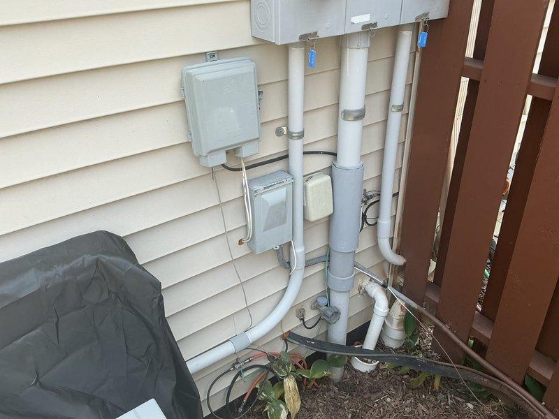 Conducted on-site assessment of 22-year-old HVAC system in condo unit to provide replacement estimate. Inspected exterior equipment location, documented existing condensing unit placement adjacent to wooden fence line, and photographed current installation configuration including refrigerant line routing, electrical service connections, and drainage piping. Evaluated existing PVC condensate drain lines, electrical disconnect box, and conduit runs mounted on exterior wall to determine compatibility with replacement equipment specifications and identify any necessary modifications for new system installation.