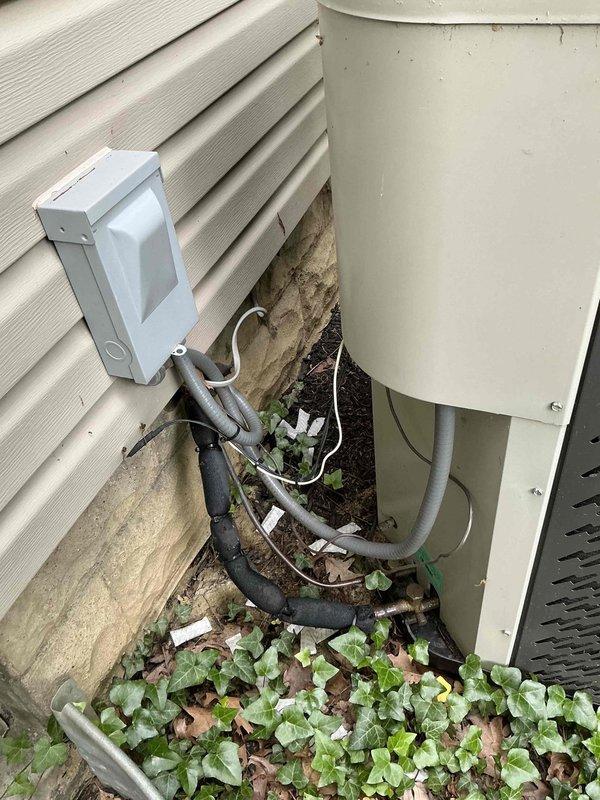 Completed heat pump replacement estimate for residential property with beige vinyl siding. Assessed existing outdoor HVAC installation including cylindrical heat pump unit positioned against exterior wall with refrigerant lines running to white electrical disconnect box mounted on foundation. Documented current system configuration showing insulated refrigerant lines, weatherproof disconnect enclosure, and unit placement on ground with surrounding vegetation. Provided replacement options and specifications for new heat pump system installation with pull-straight configuration toward house structure.