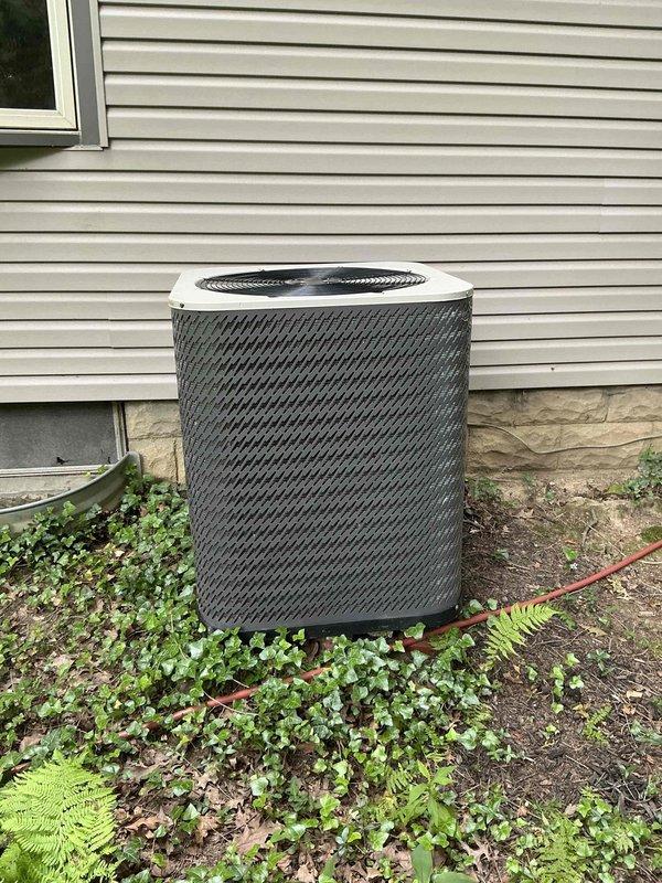 Completed heat pump replacement estimate for residential property with beige vinyl siding. Assessed existing outdoor HVAC installation including cylindrical heat pump unit positioned against exterior wall with refrigerant lines running to white electrical disconnect box mounted on foundation. Documented current system configuration showing insulated refrigerant lines, weatherproof disconnect enclosure, and unit placement on ground with surrounding vegetation. Provided replacement options and specifications for new heat pump system installation with pull-straight configuration toward house structure.
