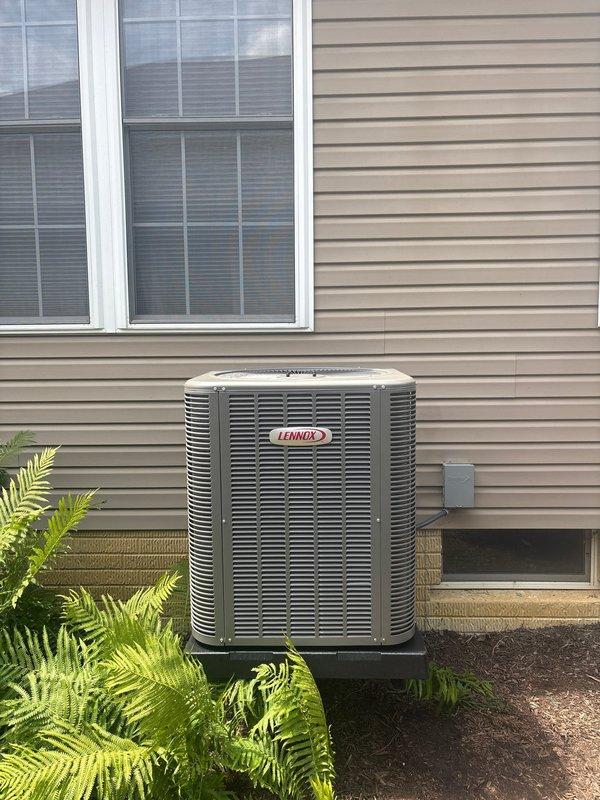 Completed installation of new Lennox air conditioning condensing unit with matching evaporator coil. Unit was properly positioned on elevated platform stand against exterior wall with adequate clearance for airflow and service access. Electrical connections were established including installation of new fuses and disconnect components to meet code requirements. System was tested for proper operation including fan motor function and electrical continuity. Unit installation includes professional landscaping consideration with existing ferns maintained around equipment location.