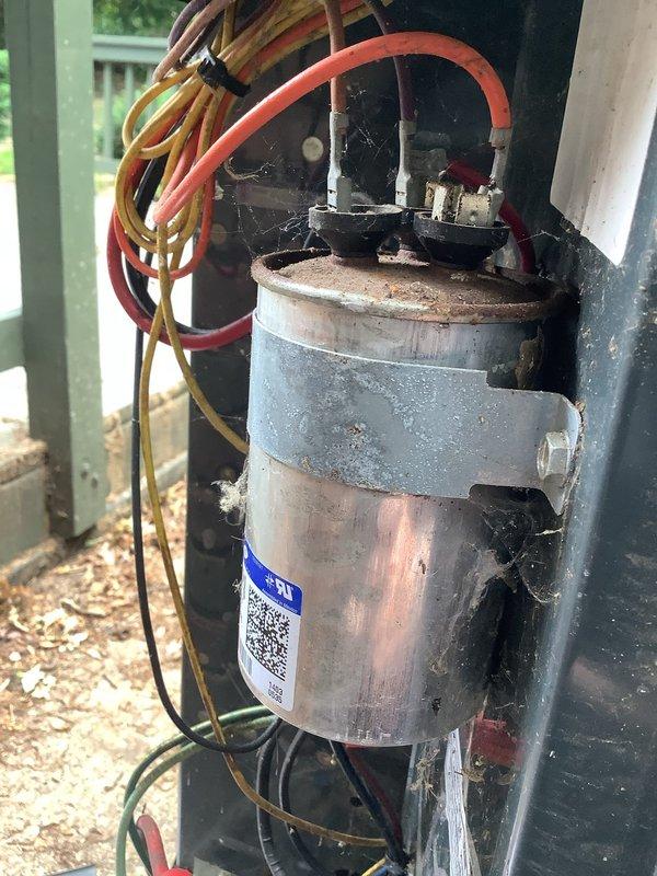 # HVAC Service Completed

Responded to service call for Trane central air conditioning system with inadequate cooling performance. Customer recently moved into property and reported unit not pumping cold air effectively, with unknown equipment age and service history.

Performed comprehensive system inspection of outdoor condenser unit. Conducted visual assessment revealing aged capacitor with significant weathering, discoloration, and corrosion on exterior casing. Identified deteriorated duct tape wrapping around capacitor body and presence of cobwebs near terminal connections, indicating extended period without maintenance. Examined electrical connections at capacitor terminals, noting white ceramic insulators and multiple wire connections including orange, red, and black leads.

Inspected condenser unit exterior condition, observing dirt accumulation on top surface and sides, with debris collection around base and foundation area. Verified fan grille integrity and overall structural
