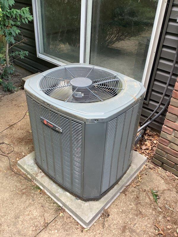 # HVAC Service Completed

Responded to service call for Trane central air conditioning system with inadequate cooling performance. Customer recently moved into property and reported unit not pumping cold air effectively, with unknown equipment age and service history.

Performed comprehensive system inspection of outdoor condenser unit. Conducted visual assessment revealing aged capacitor with significant weathering, discoloration, and corrosion on exterior casing. Identified deteriorated duct tape wrapping around capacitor body and presence of cobwebs near terminal connections, indicating extended period without maintenance. Examined electrical connections at capacitor terminals, noting white ceramic insulators and multiple wire connections including orange, red, and black leads.

Inspected condenser unit exterior condition, observing dirt accumulation on top surface and sides, with debris collection around base and foundation area. Verified fan grille integrity and overall structural