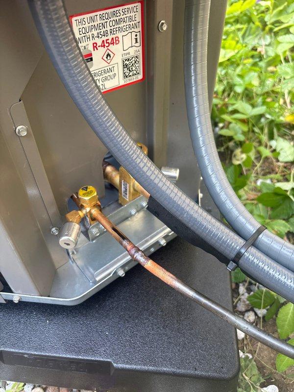 Completed installation of outdoor air conditioning condenser unit with R-454B refrigerant system. Unit was properly mounted on platform base against exterior wall with electrical disconnect box and conduit installed per code requirements. Refrigerant lines were connected to brass valve assembly and routed through unit, with all connections secured and tested for proper operation.