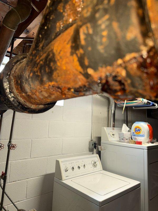 Responded to service call for cracked pipe in basement. Upon inspection, discovered severely corroded and deteriorated metal piping system in utility room area above washing machine. The existing pipe exhibited extensive rust, oxidation, and structural compromise throughout, with flaking metal and significant corrosion damage that created failure points in the system. Documented conditions with photographs showing the extent of deterioration to both the main cylindrical ductwork/piping and associated plumbing infrastructure mounted to ceiling joists. Assessed surrounding copper, PVC, and metal pipe connections, valves, and mounting hardware for related damage. Provided estimates for pipe replacement and repair options to address the compromised plumbing system.