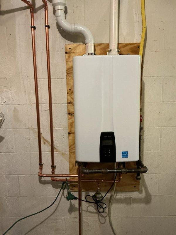 Completed full replacement of wall-mounted Navien tankless water heater unit in residential basement utility area. Removed existing unit and installed new tankless water heater on plywood backing board against cinderblock wall, connecting all copper water supply lines, gas lines, electrical wiring, and white PVC exhaust venting. Installation included proper mounting, connection of all utilities, and verification of operational status with digital display panel functioning correctly.