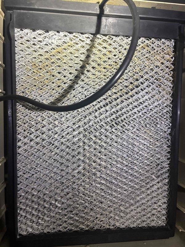 Performed routine maintenance inspection on residential air conditioning system. Examined indoor evaporator coil and filter assembly, noting diamond-patterned mesh filter in proper working condition with drainage line secured. Inspected outdoor condenser unit on concrete pad, observing weathered heat exchanger coils with accumulated environmental debris typical of outdoor exposure. System operational at time of service.