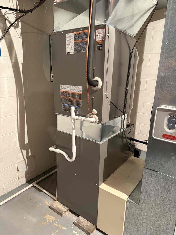 Completed installation of 97% efficiency furnace in residential basement utility area. Previous furnace failure had resulted in loss of cooling capability for the recently installed air conditioning system. Installed new gray metal forced-air furnace unit with proper venting configuration using white PVC intake and exhaust piping extending vertically from unit top with elbow fittings. Secured all electrical connections with black wiring routed to upper section of installation. Connected metal supply and return ductwork to furnace air handler. Installed condensate drainage line using white PVC piping at lower unit section. Verified proper integration with existing air conditioning equipment and water heater in shared mechanical space. All connections secured per manufacturer specifications and local code requirements. System tested and operational.
