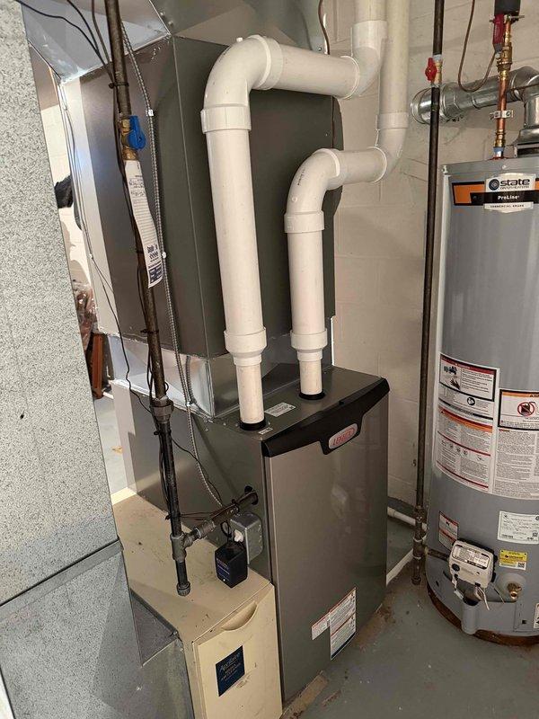 Completed installation of 97% efficiency furnace in residential basement utility area. Previous furnace failure had resulted in loss of cooling capability for the recently installed air conditioning system. Installed new gray metal forced-air furnace unit with proper venting configuration using white PVC intake and exhaust piping extending vertically from unit top with elbow fittings. Secured all electrical connections with black wiring routed to upper section of installation. Connected metal supply and return ductwork to furnace air handler. Installed condensate drainage line using white PVC piping at lower unit section. Verified proper integration with existing air conditioning equipment and water heater in shared mechanical space. All connections secured per manufacturer specifications and local code requirements. System tested and operational.