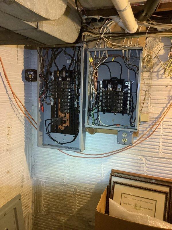 Responded to service call for downed electrical line between main residence and detached garage caused by fallen tree branch. Upon arrival, confirmed customer had properly de-energized the circuit at the breaker panel as a safety precaution. Assessed the overhead service conductors running from house to garage, documenting damage to the aerial span and connection points. Photographed the garage interior showing the automatic door opener system and electrical infrastructure, as well as the dual breaker panel configuration in the utility space. Verified proper breaker positioning and panel condition to ensure safe isolation of the affected circuit serving the garage structure.