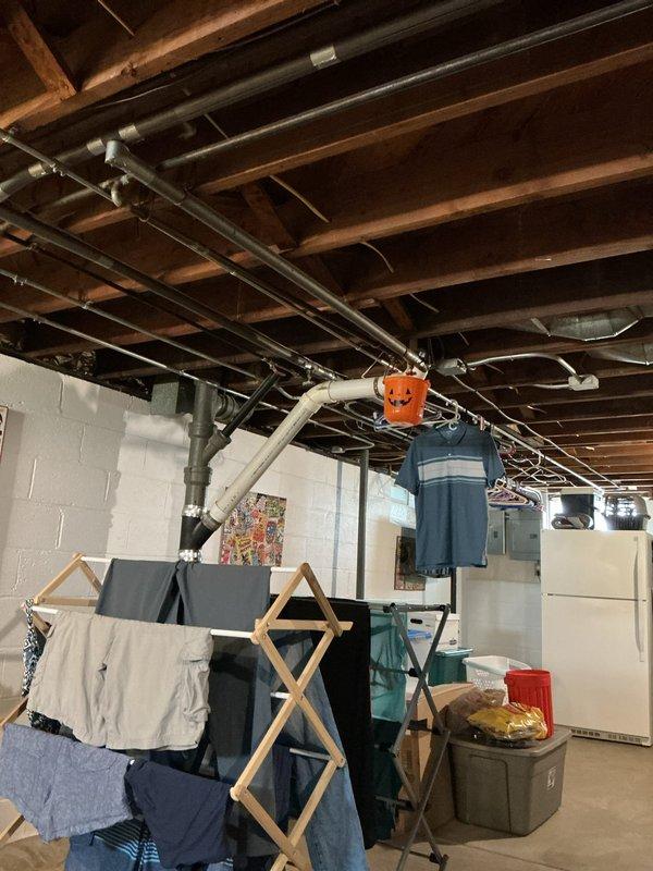 Responded to customer report of thumping noise behind wall when faucet was running and slow drip from pipe traveling downstairs with unknown source. Upon inspection, located active leak in overhead plumbing system in basement area. Positioned collection bucket beneath leaking pipe to prevent water damage while assessing the issue. Inspected exposed PVC piping, connections, and fittings throughout the basement ceiling joists to identify the source of both the audible noise and visible water leak.