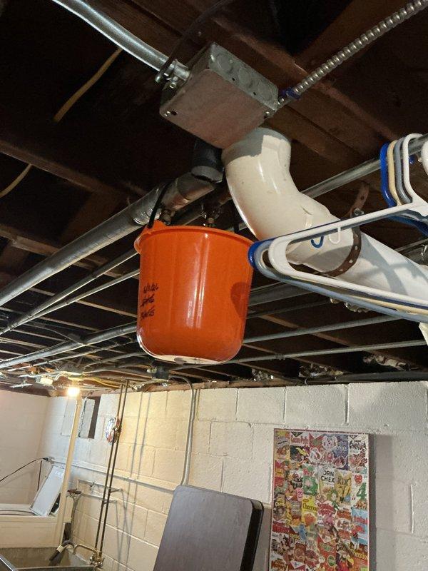 Responded to customer report of thumping noise behind wall when faucet was running and slow drip from pipe traveling downstairs with unknown source. Upon inspection, located active leak in overhead plumbing system in basement area. Positioned collection bucket beneath leaking pipe to prevent water damage while assessing the issue. Inspected exposed PVC piping, connections, and fittings throughout the basement ceiling joists to identify the source of both the audible noise and visible water leak.