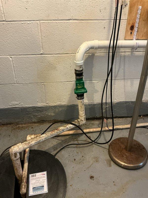 Responded to callback for leaking sump pump discharge line that was recently replaced. Upon inspection, identified leak at connection point between white PVC discharge pipe and check valve assembly. Found deteriorated copper piping and corroded components in the discharge system contributing to improper sealing. Evaluated the PVC elbow joint, black coupling connection, and green check valve for proper installation and integrity. Documented existing conditions including aged copper horizontal discharge line showing significant oxidation and patina buildup along floor level, mixed old and new plumbing components, and overall system configuration. Assessed discharge assembly mounting against concrete block wall and verified proper slope and support of discharge piping to prevent future leakage issues.