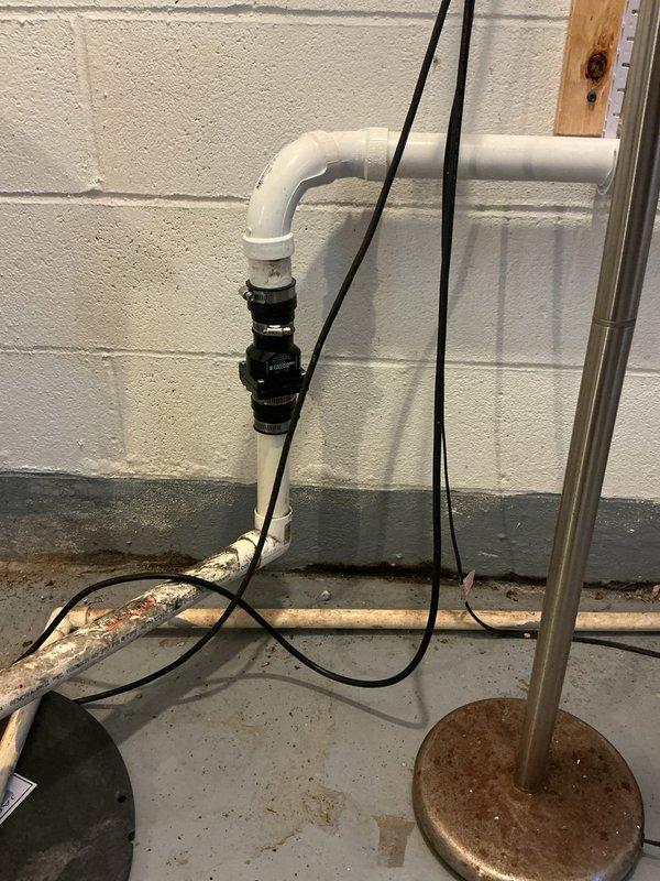 Responded to callback for leaking sump pump discharge line that was recently replaced. Upon inspection, identified leak at connection point between white PVC discharge pipe and check valve assembly. Found deteriorated copper piping and corroded components in the discharge system contributing to improper sealing. Evaluated the PVC elbow joint, black coupling connection, and green check valve for proper installation and integrity. Documented existing conditions including aged copper horizontal discharge line showing significant oxidation and patina buildup along floor level, mixed old and new plumbing components, and overall system configuration. Assessed discharge assembly mounting against concrete block wall and verified proper slope and support of discharge piping to prevent future leakage issues.