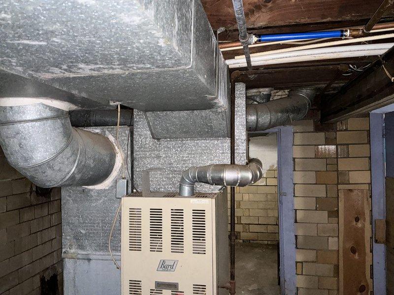 Conducted on-site assessment for furnace and air conditioning replacement estimate. Documented existing HVAC system including heavily dust-laden air handler unit and Bard furnace located in basement utility area. Evaluated deteriorated ductwork installation with significant structural damage to concrete block wall at penetration point, including crumbled and missing blocks requiring repair. Photographed aged equipment showing extended period without maintenance, compromised duct connections, and overall system condition to support replacement recommendations for complete heating and cooling system upgrade.