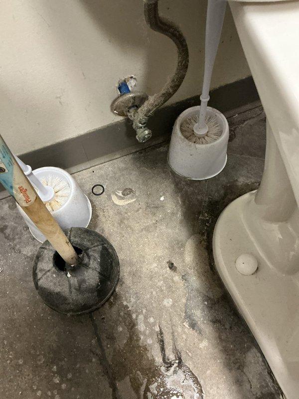 Responded to water leak at baseboard near toilet in men's restroom. Upon inspection, identified source of moisture infiltration affecting floor area around toilet base in facility with 10-year-old plumbing system. Assessed water supply line connections, shut-off valve, and wax ring seal integrity. Documented existing floor staining and water damage patterns on concrete surface. Photographed plumbing infrastructure and fixture condition for service records.